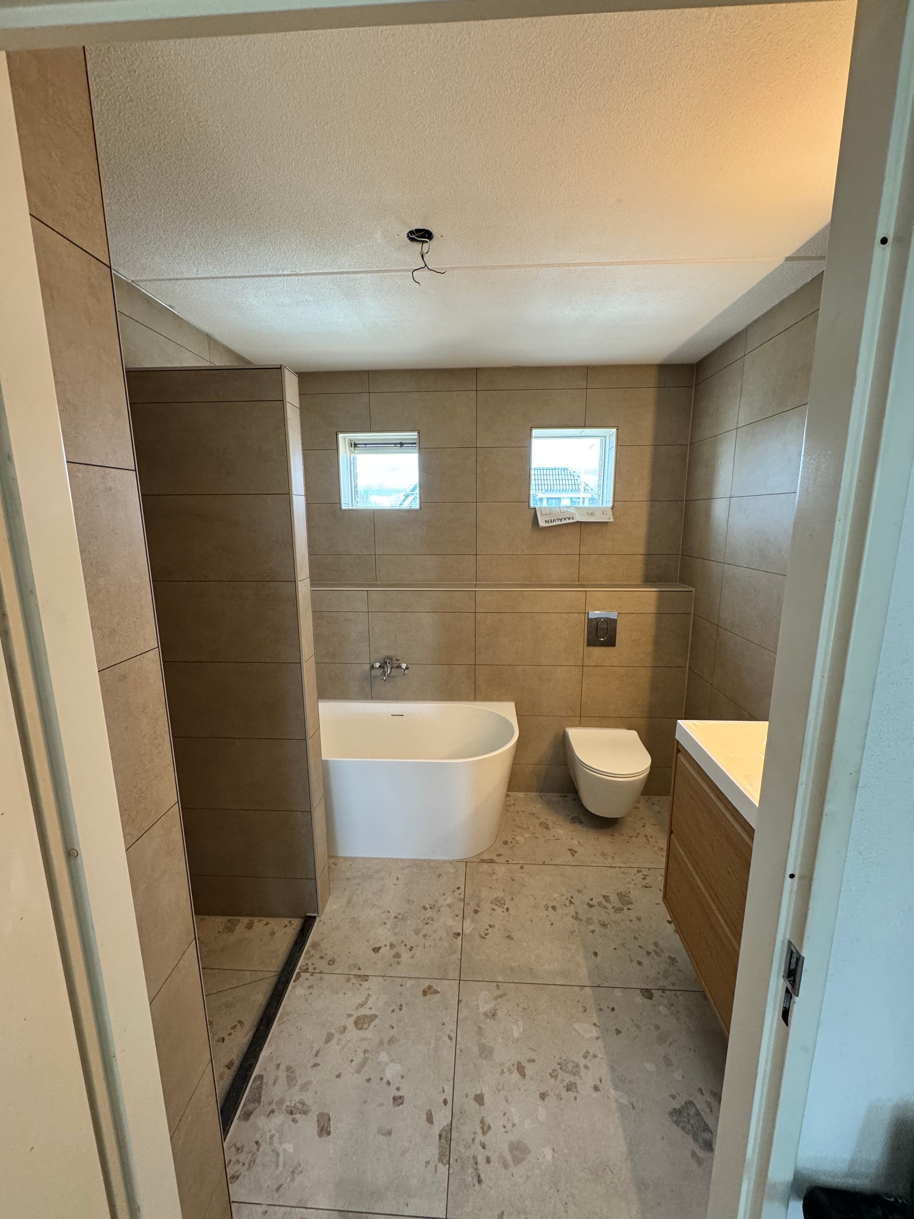 Moderne badkamer met beige wandtegels, wit vrijstaand bad, hangend toilet en houten wastafelmeubel.