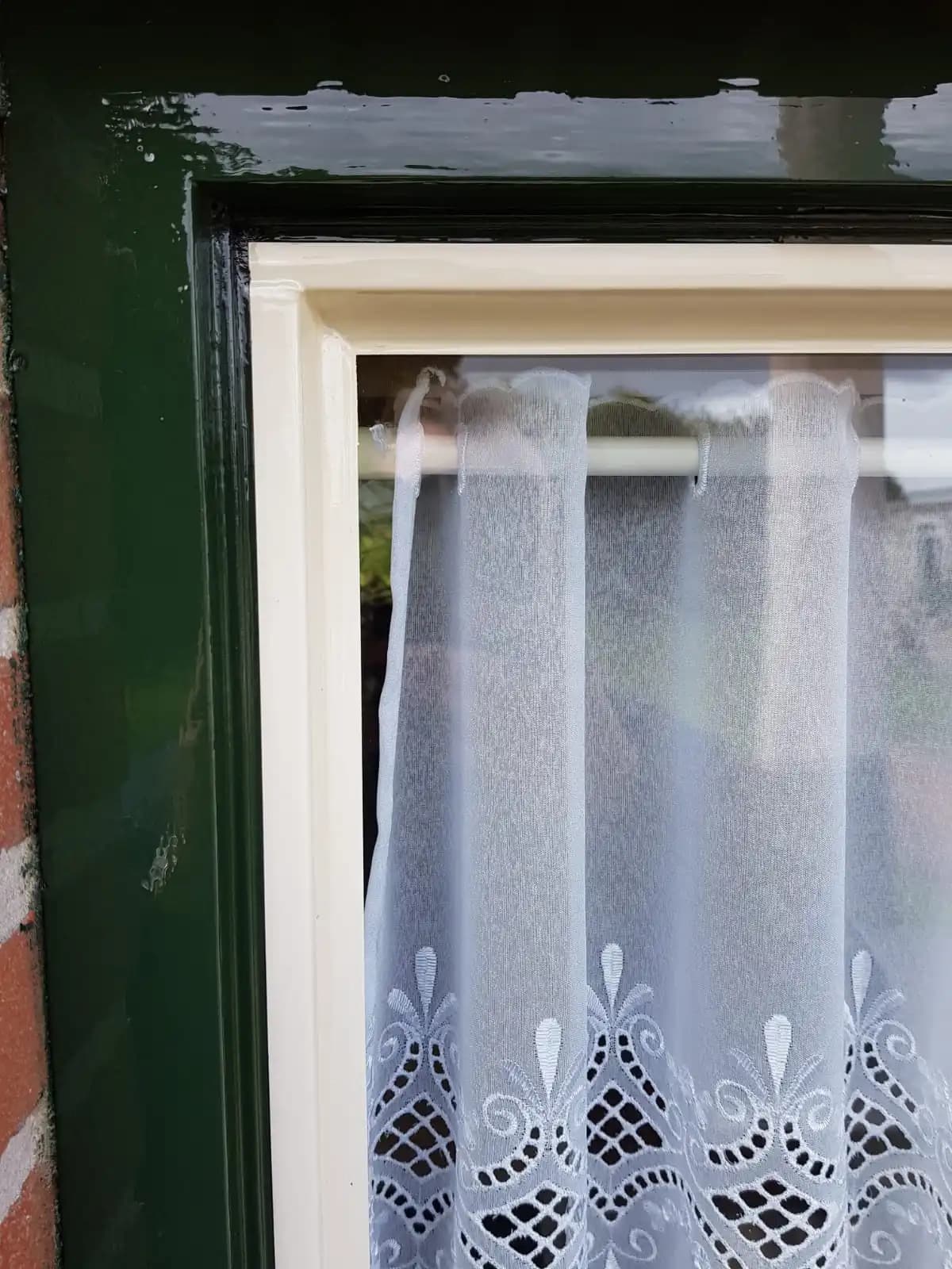 Wit kanten gordijn hangt voor een raam met een donkergroen en crème-kleurig kozijn.
