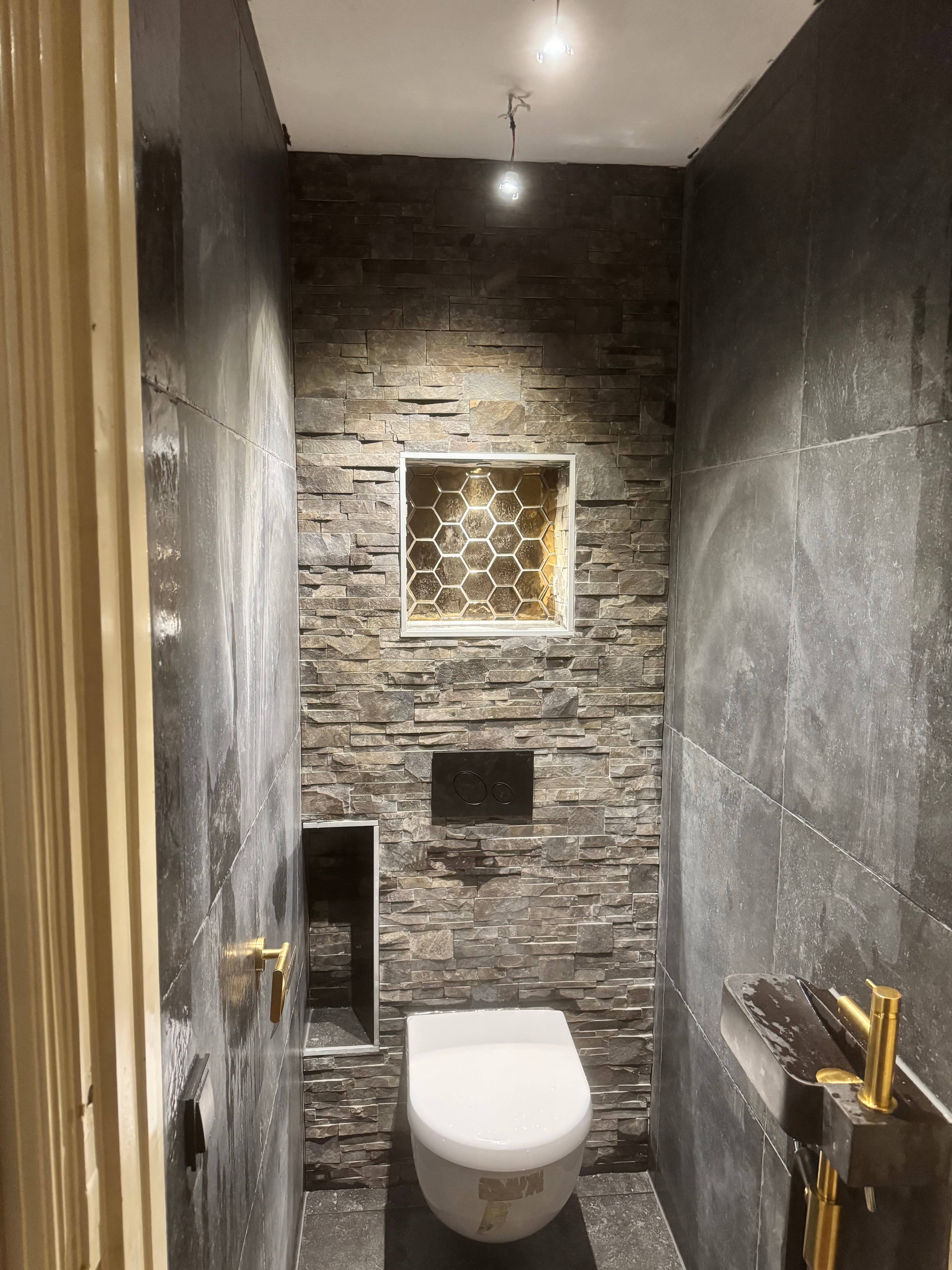 Moderne toiletruimte met stenen achterwand, wit toilet, gouden accenten en een verlichte honingraat nis.