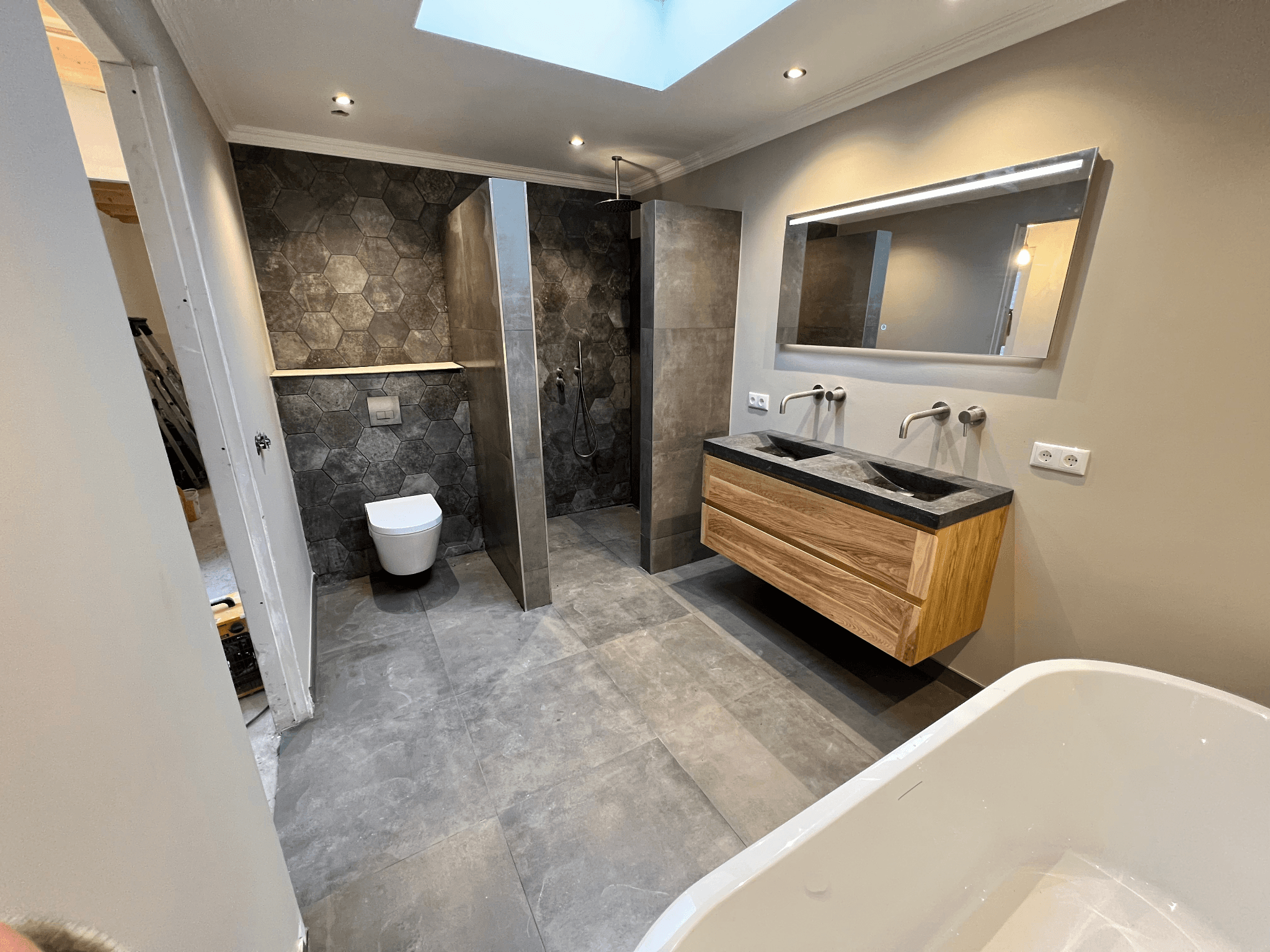 Moderne badkamer met grijze tegels, hexagonale wanden, houten wastafelmeubel, dubbele wastafels, inloopdouche en toilet.