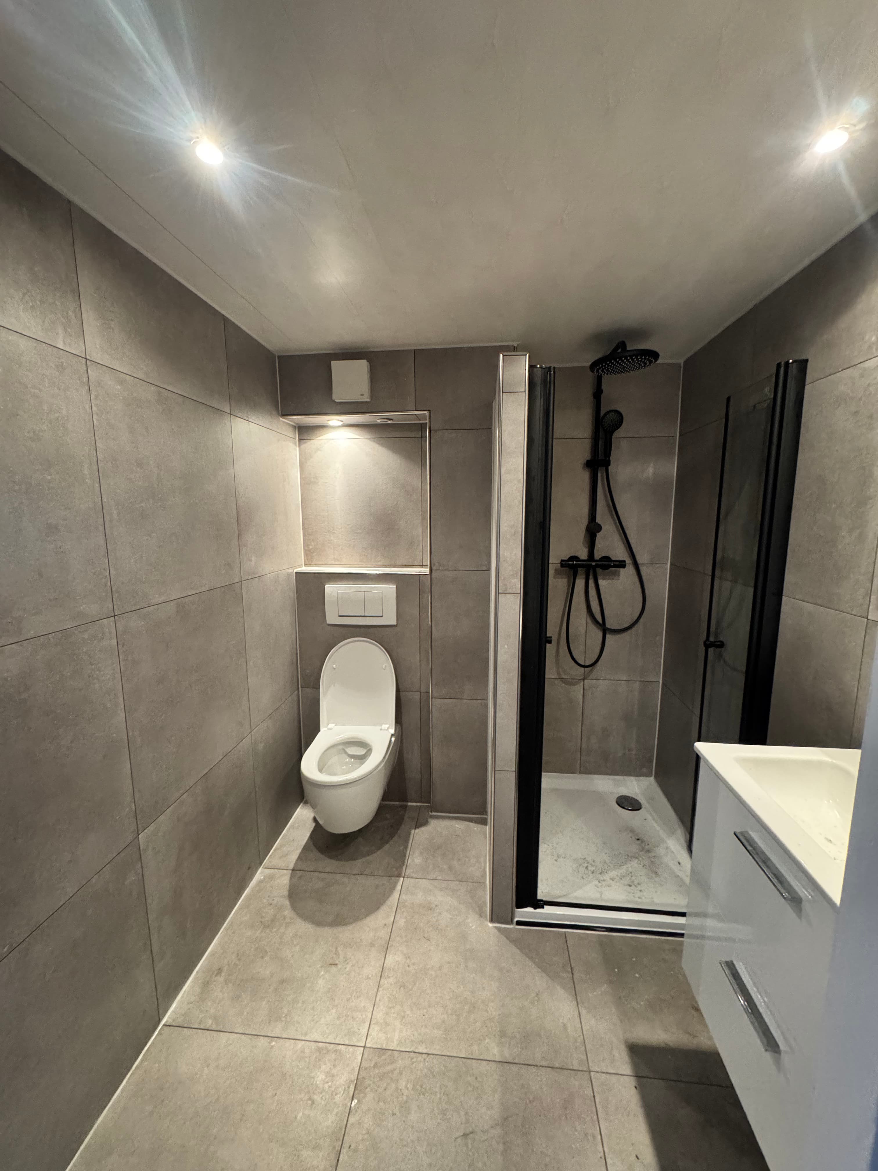 Moderne badkamer met grijze tegels, wit toilet, inloopdouche met zwarte armaturen en wit wastafelmeubel.